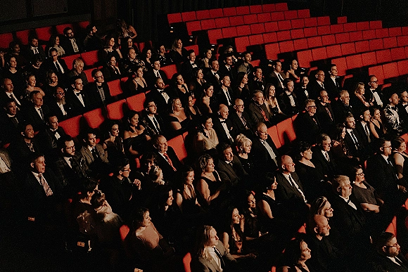Wedding guests in formal attire seated in red theater chairs, tuxedos and dresses visible in a dark auditorium venue interior