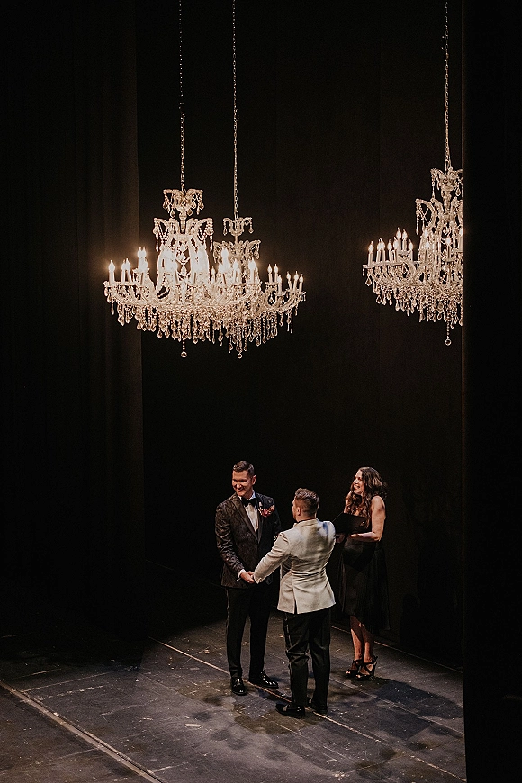 Wedding vows as two grooms hold hands during a same sex wedding ceremony under crystal chandeliers and candlelight on a dark stage