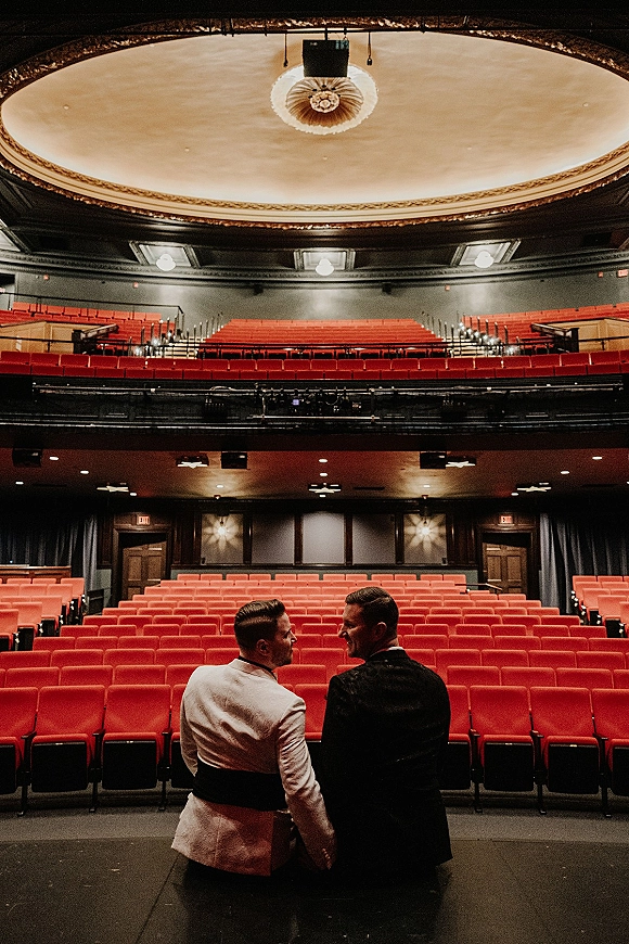 Wedding couple portrait of two grooms holding hands, one in a white dinner jacket, looking back in an ornate red-seat theater auditorium
