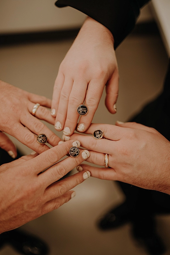 Wedding ring close-up of stacked hands showing wedding bands, matching signet rings, manicured nails, and black suit sleeves on a blurred floor