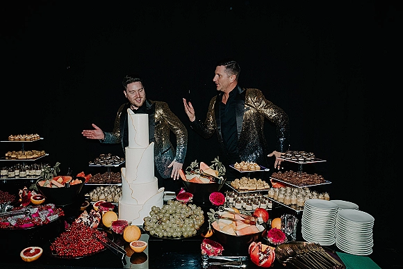Wedding dessert table with tiered white wedding cake, dessert stands of mini sweets and fruit platters, set against a black backdrop