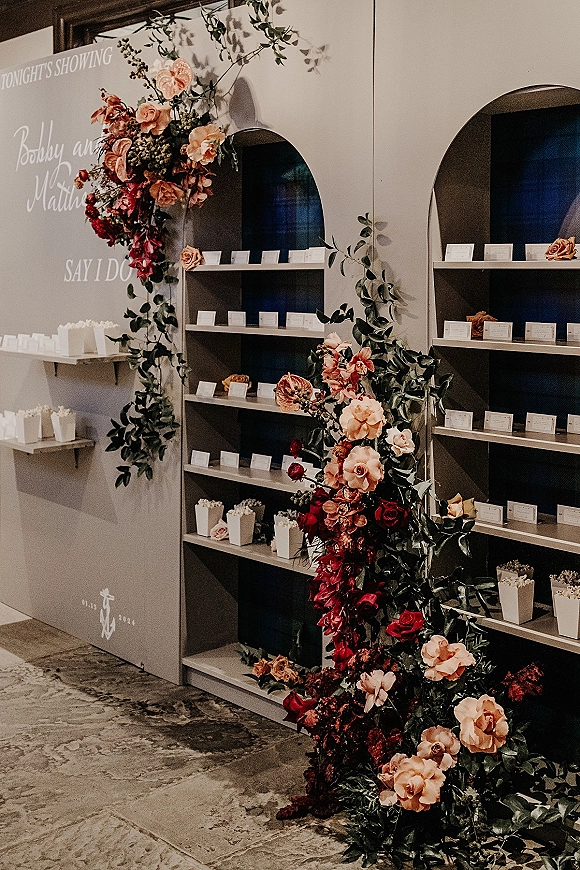 Escort card display on built-in shelves with a rose and greenery garland accent, place cards and popcorn boxes against arched niches