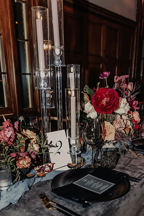 Reception tablescape with a wedding centerpiece with candles, black plates and gold flatware on a gray velvet tablecloth by dark wood walls