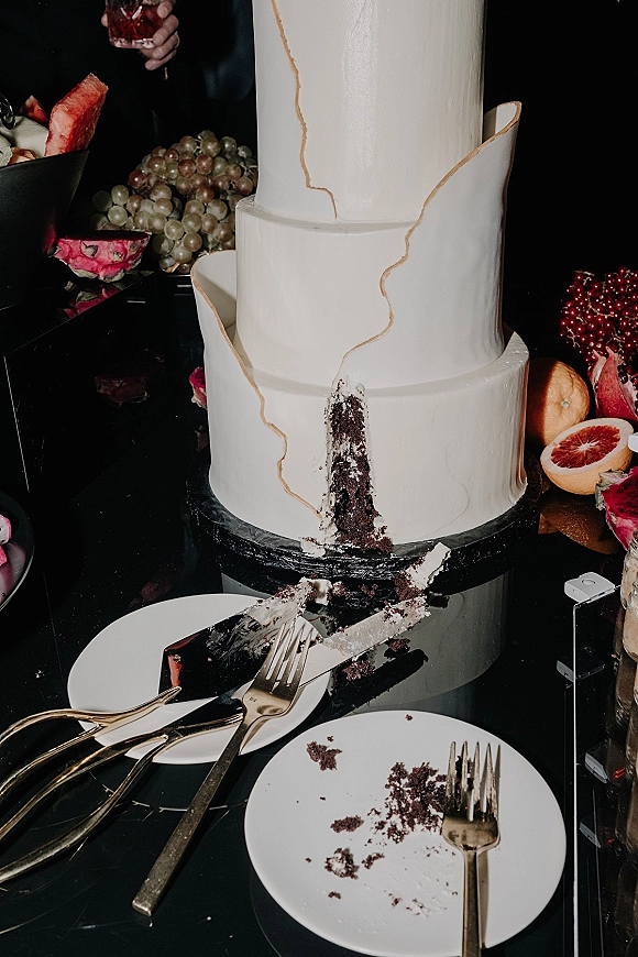 Wedding cake with gold piping on a stand, slice removed with crumbs, fruit styling and gold cutlery on a dark table backdrop