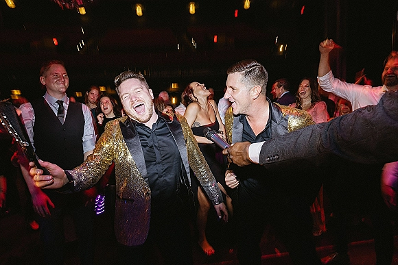 Wedding reception party with guests dancing as a man in a gold sequin dinner jacket sings into a handheld microphone under string lights