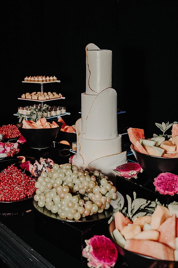 Wedding cake with gold detailing on a dessert stand, surrounded by grapes, melon slices and dragon fruit on a black dessert table backdrop
