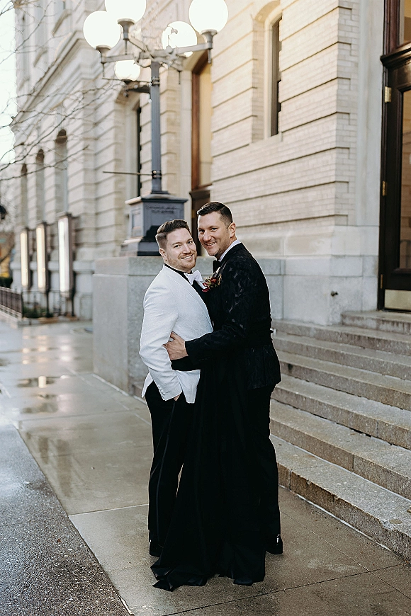 Couple portrait of two grooms embracing in tuxedos, one in a white dinner jacket, on wet steps outside a stone building