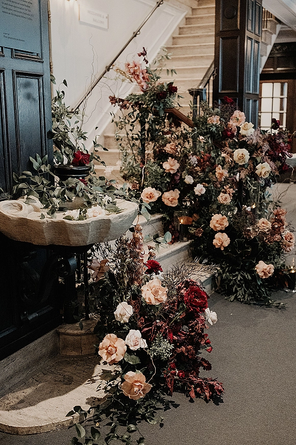 Wedding floral decor cascades along an interior staircase floral installation with roses, greenery garland, and candlelit stone pedestals