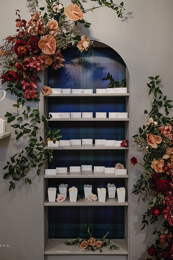 Seating chart display with escort card display on built-in shelves, greenery garland and anthurium blooms in an arched niche with blue backdrop