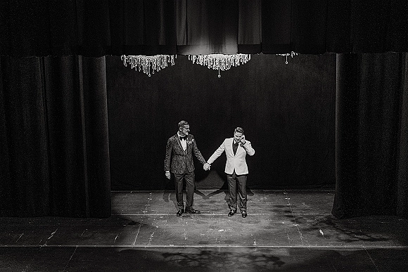 Groom portrait in a classic tuxedo with bow tie and boutonniere, standing on a theater stage before black curtains under chandelier light