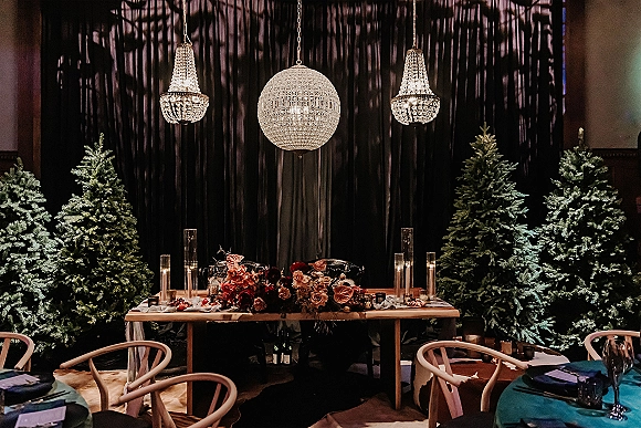 Sweetheart table decor with taper candles and a low floral centerpiece beneath crystal chandeliers and a disco ball against a dark velvet backdrop