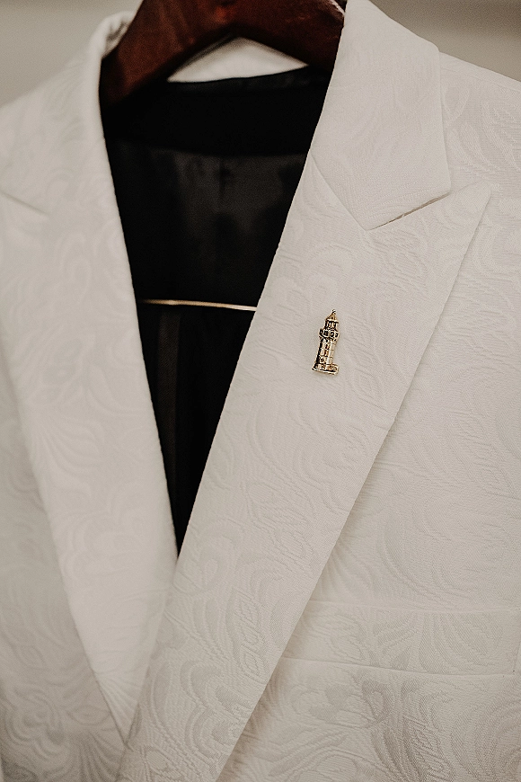 Groom suit detail featuring a white tuxedo jacket with black bow tie, crisp dress shirt, and lapel pin against an indoor backdrop