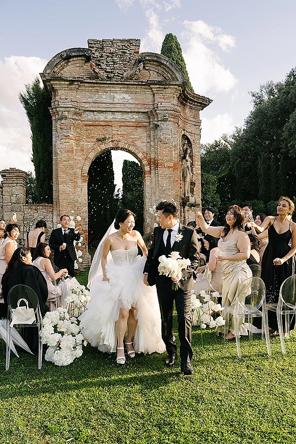 Wedding recessional with ceremony exit confetti as bride and groom walk the aisle, guests cheering beneath a stone archway in garden ruins