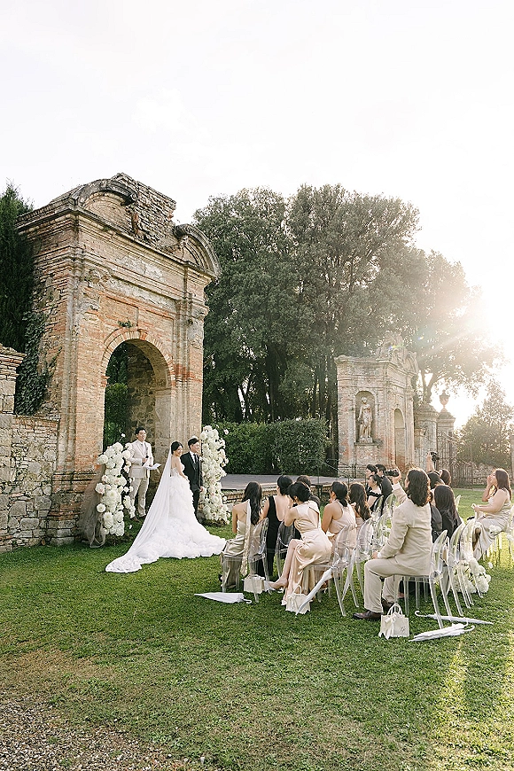 Wedding ceremony with bride and groom at altar beneath a white floral arch, clear chairs lining aisle runner at sunlit stone ruins