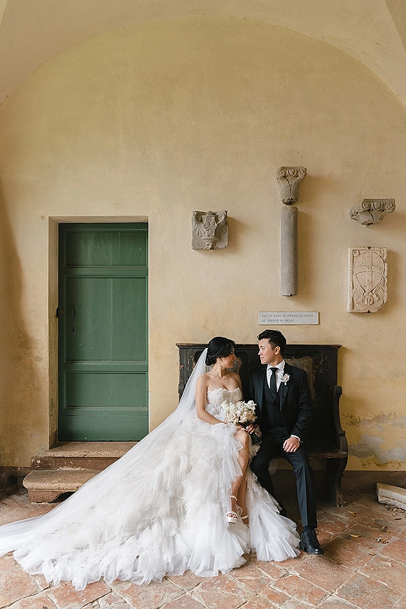 Couple portrait of bride and groom seated on a bench, bride in strapless gown with long veil holding a white orchid bouquet by a green door