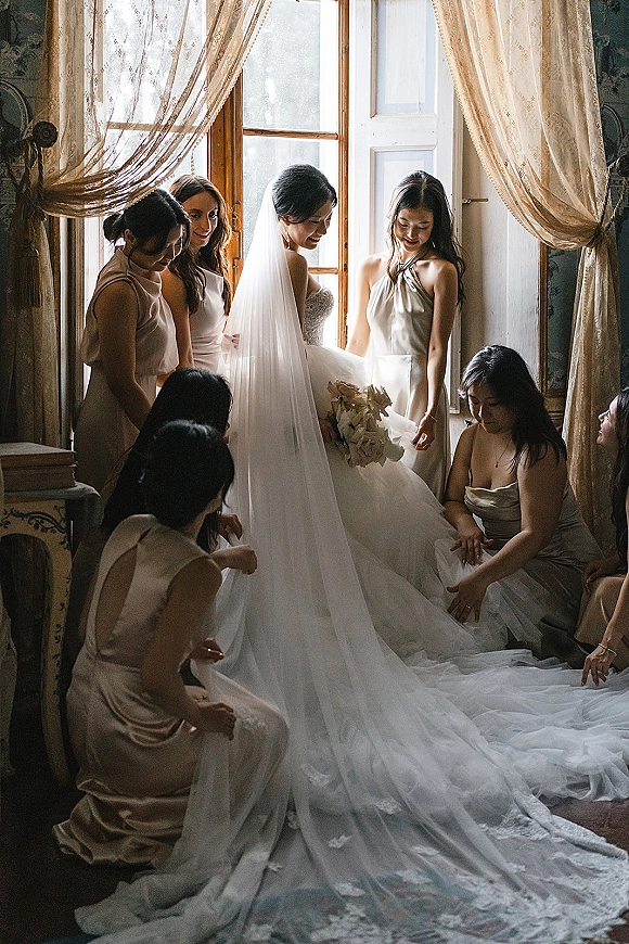 Bride getting ready as bridesmaids help adjust her long veil in a vintage room with window light and sheer curtains, holding a white bouquet