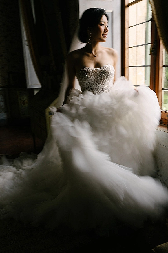 Bridal portrait of a bride by window light in a strapless beaded bodice and tulle skirt, wearing statement earrings and a pendant necklace
