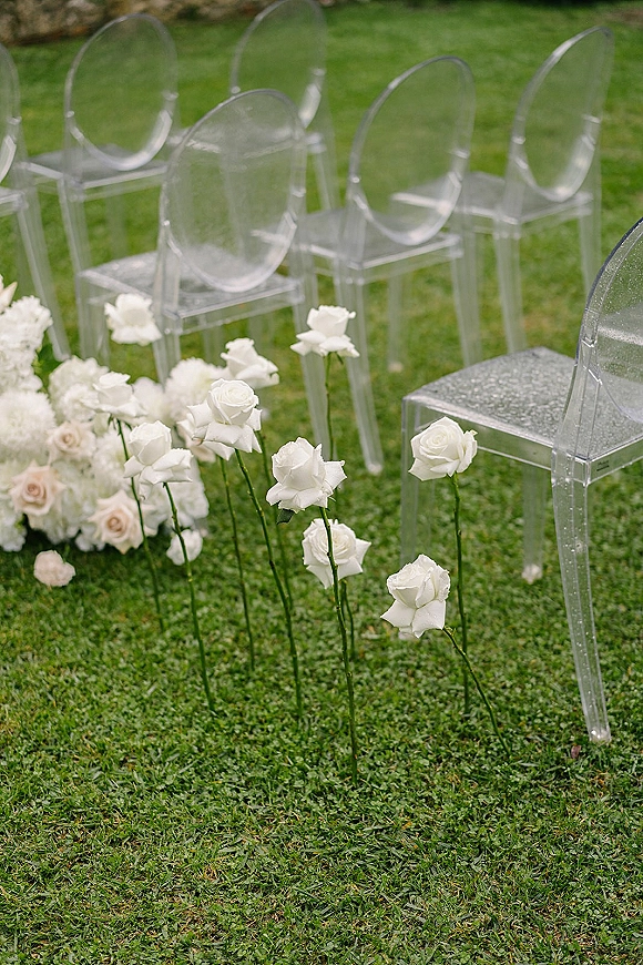 Ceremony aisle decor with clear acrylic chairs lined on a green lawn, accented by white and blush roses and white hydrangea clusters