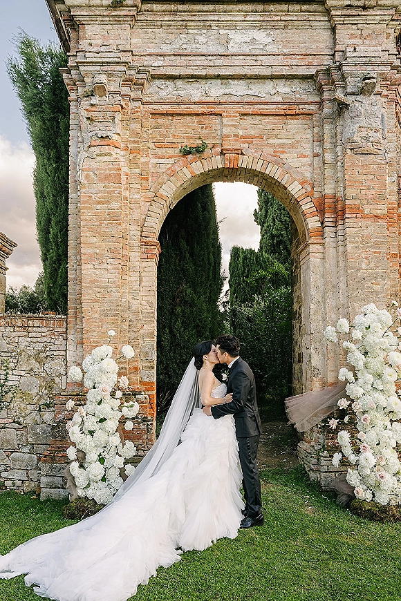 Wedding kiss portrait of bride and groom kissing beneath a stone and brick archway, bride’s long veil and train flowing by blush florals