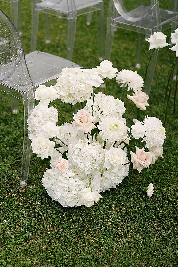 Ceremony aisle flowers with white hydrangeas and roses arranged on the grass lawn beside clear acrylic chairs for outdoor vows