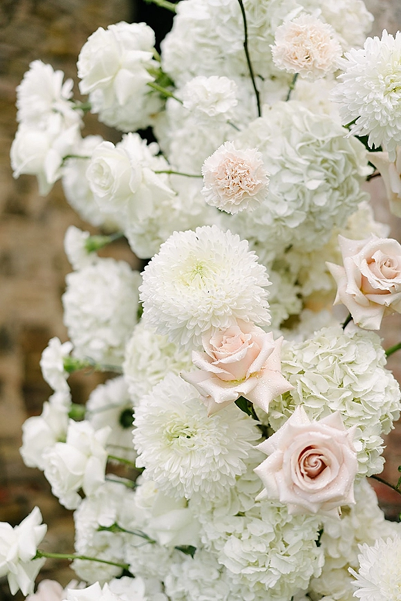 Wedding flowers with white blooms and blush roses, hydrangea, carnations, and chrysanthemums arranged against a stone wall backdrop
