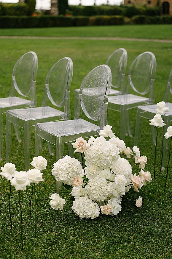 Ceremony aisle decor with clear acrylic ceremony chairs lined by white and blush hydrangea and rose arrangements on a garden lawn near a stone building