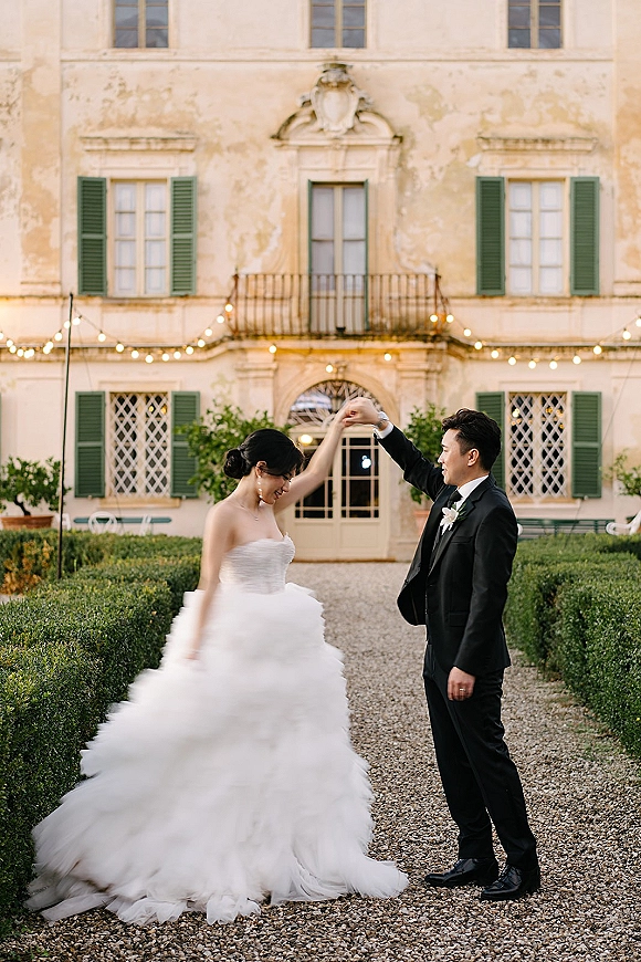 Couple portrait of bride twirling her strapless tulle ball gown as groom in tux watches under string lights in a villa courtyard