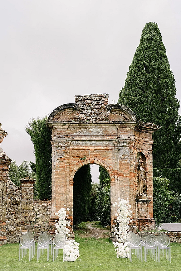 Ceremony setup for outdoor wedding ceremony with clear acrylic chairs and white floral aisle arrangements leading to a stone arch in brick ruins