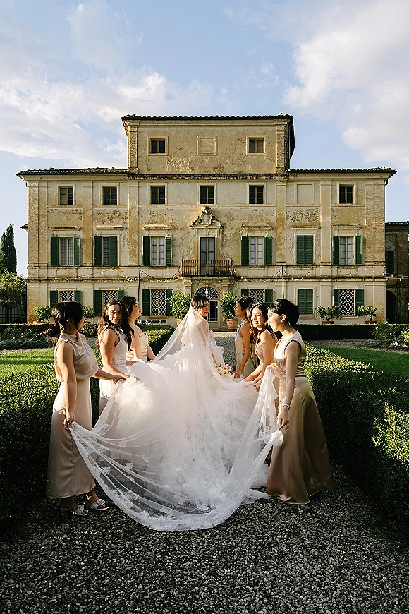 Bridal party portrait of bride with bridesmaids holding veil, lace train flowing on a gravel path before a villa with string lights and hedges