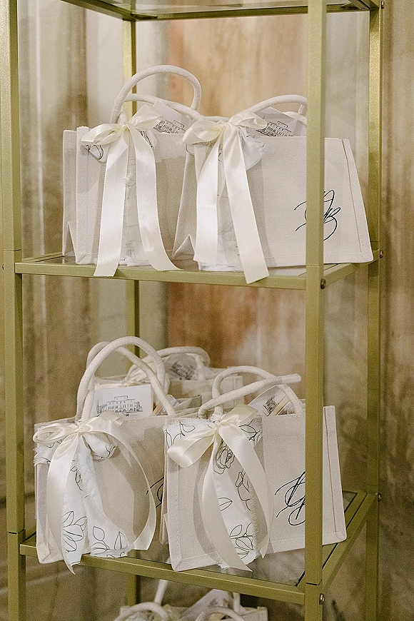 Wedding welcome bags lined on a gold shelf, white monogram tote bags with satin bow accents, tissue paper and printed cards against marble wall
