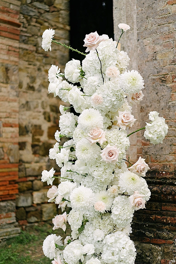 Wedding floral arrangement with hydrangea rose blooms, white dahlias, carnations, and greenery cascading beside a stone doorway on brick wall