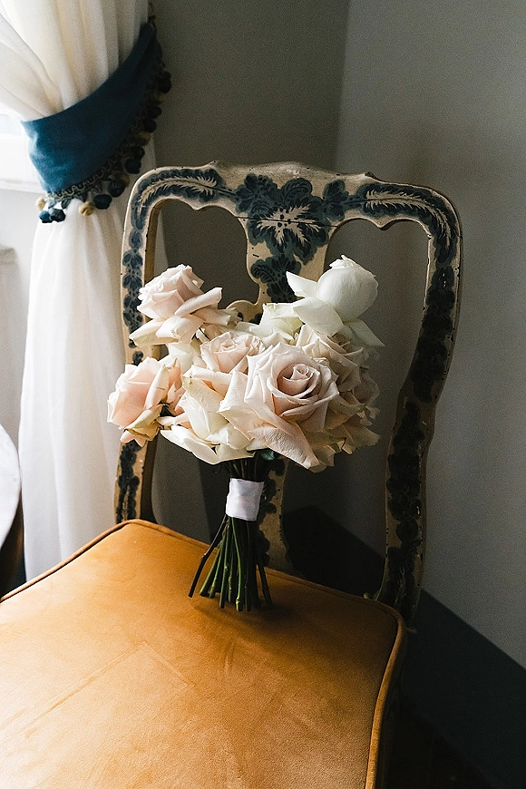Bridal bouquet of blush roses and white roses with ribbon wrap resting on a vintage chair by a gray wall in soft window light
