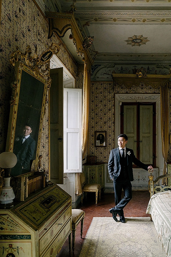 Groom portrait in black suit with tie and boutonniere, standing by an open window in a vintage bedroom with an ornate gold mirror