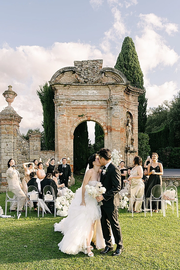 Wedding kiss as bride in strapless gown and veil kisses groom in black tux under a stone archway, holding a white floral bouquet outdoors