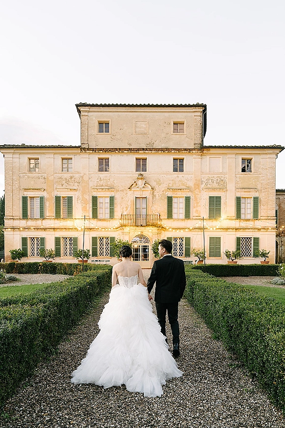 Couple portrait of bride and groom walking away hand in hand, her long train flowing on a garden path before a villa with string lights