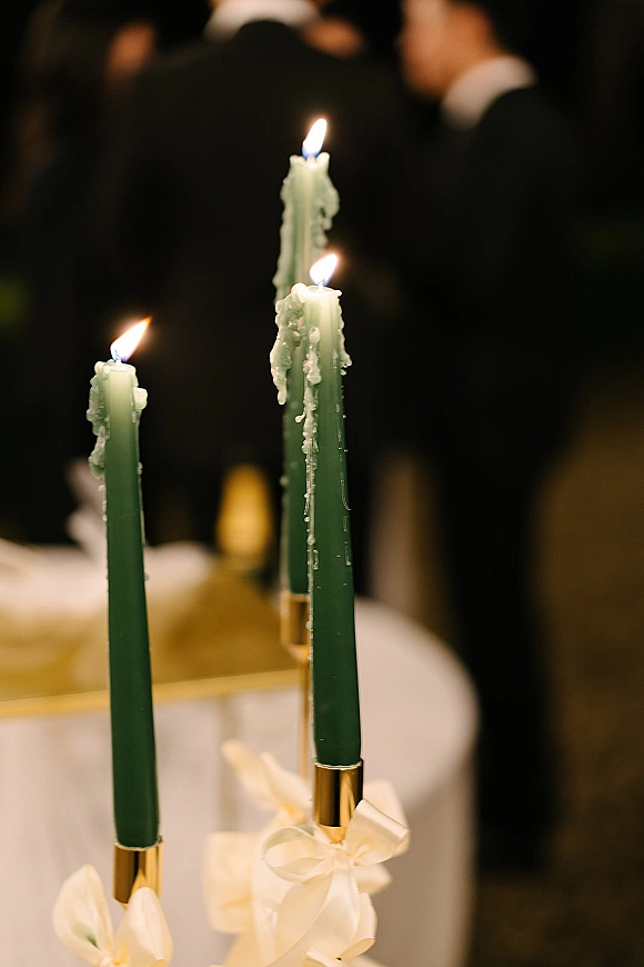 Taper candles with green taper candles drip wax in gold holders, tied with white ribbon bows on a reception table with blurred guests