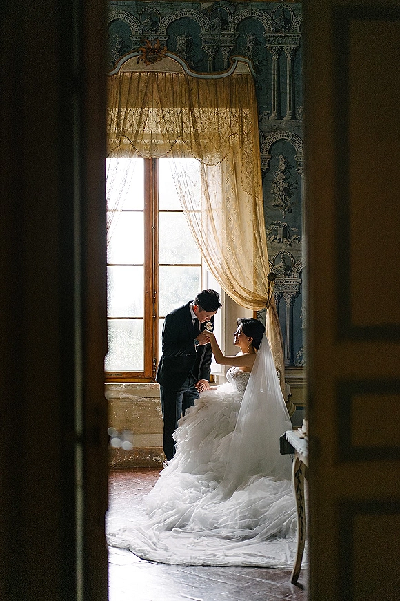 Couple portrait of groom kissing the bride’s hand by a tall window, her veil and long train flowing over wooden floor in doorway light