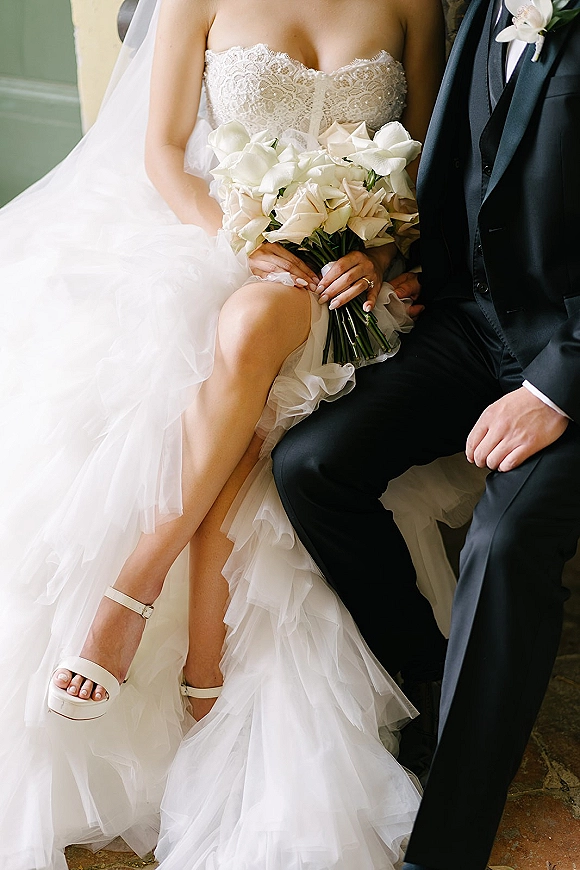 Couple portrait of bride and groom seated, bride holding a white calla lily bouquet, showing lace gown, veil, ring, and black tuxedo indoors