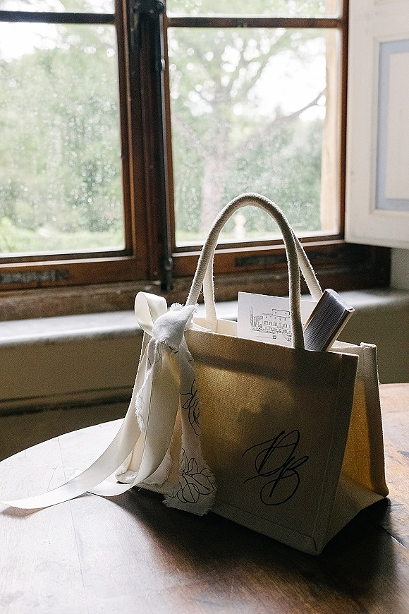 Wedding welcome bag canvas tote with monogram, white ribbon bow, welcome card and guest booklet on a wood table by a window with greenery outside