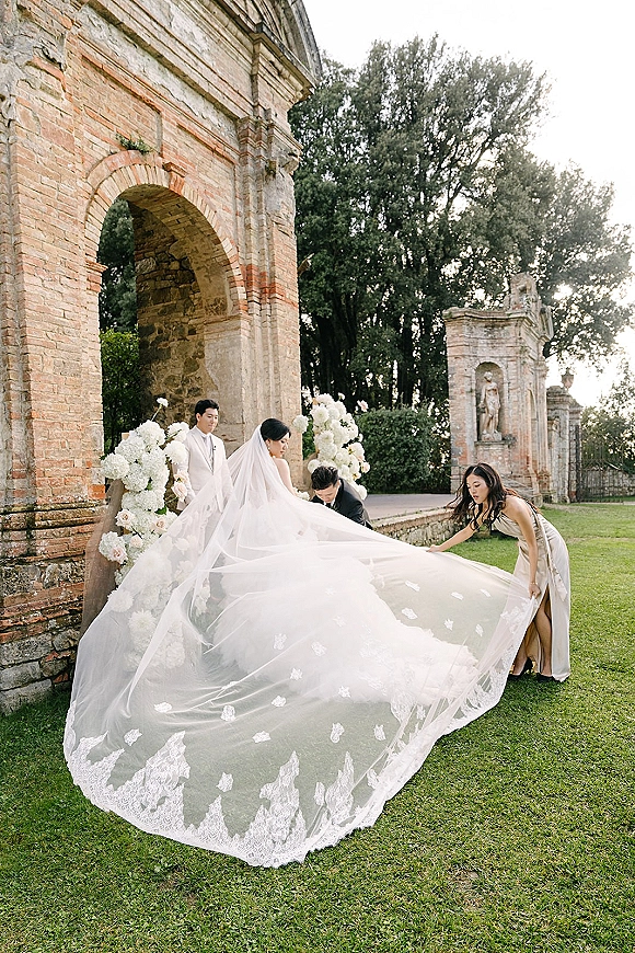 Bride portrait with a cathedral veil bride as bridesmaids adjust her lace train by a stone archway in garden ruins