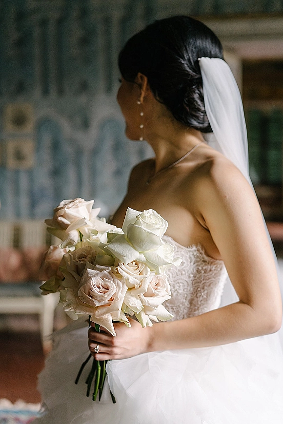 Bridal portrait of a bride holding bouquet of white and blush roses, wearing a strapless lace gown and veil in a softly lit room