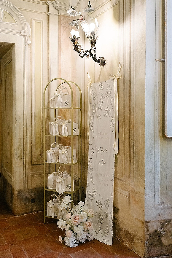 Wedding welcome sign on linen with calligraphy text, ribbon ties, and favor bags on a metal rack in a vintage hallway with tiles