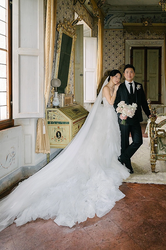 Couple portrait of bride and groom posing as she leans on him, holding a bouquet in a vintage room with ornate mirror and window light