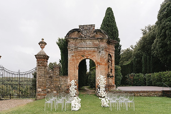 Ceremony setup with clear acrylic chairs and white floral aisle markers facing a stone archway and wrought iron gate on a garden lawn