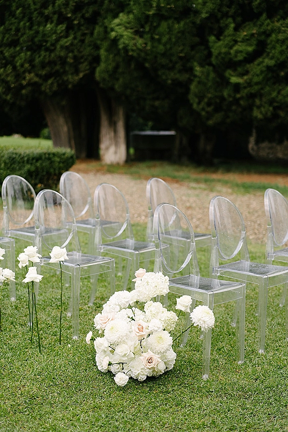 Ceremony seating with clear acrylic wedding chairs lined along an aisle, accented by white rose and hydrangea florals on a grassy lawn