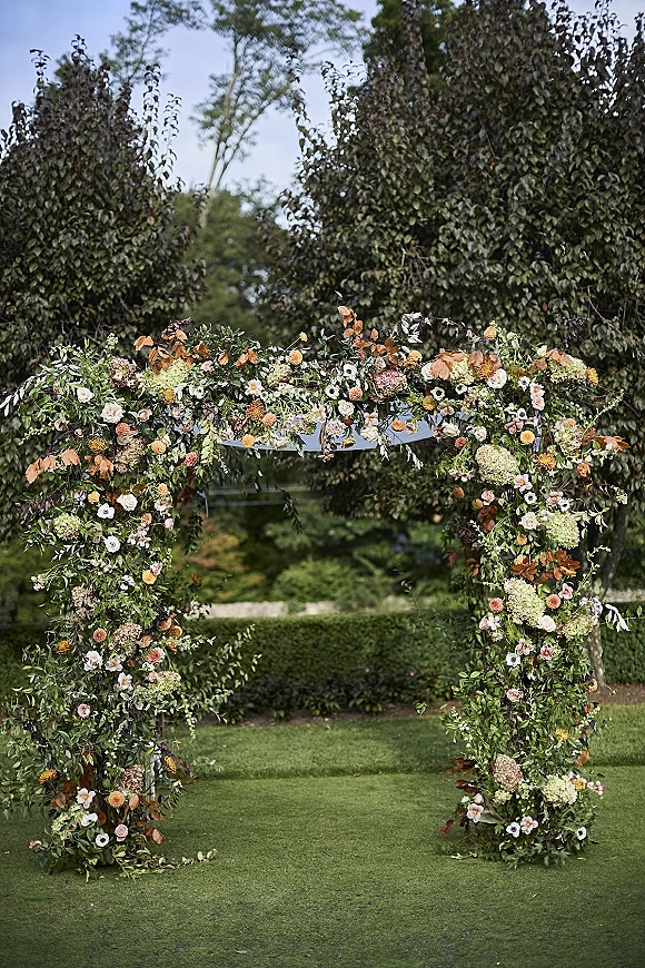 Wedding ceremony arch with a floral wedding arch of greenery garland and mixed blooms on a garden lawn with hedges and trees