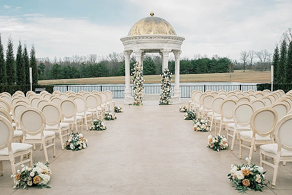 Ceremony setup for outdoor wedding ceremony with white chairs and peach-and-white floral columns framing a white gazebo by the lake
