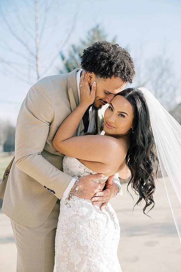 Couple portrait of bride and groom embrace, forehead touch as she looks at camera, long veil flowing outdoors with trees and sky