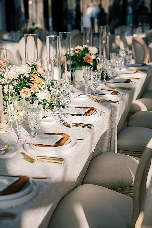 Reception tablescape with a long banquet table setup, white patterned tablecloth, rose greenery centerpieces, and floating candles in glass vases.