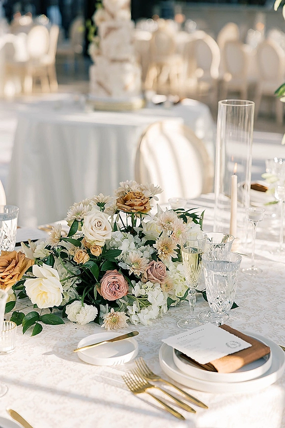 Reception tablescape with wedding table centerpiece of roses and greenery, gold flatware, crystal goblets, and candlelit glow near cake table